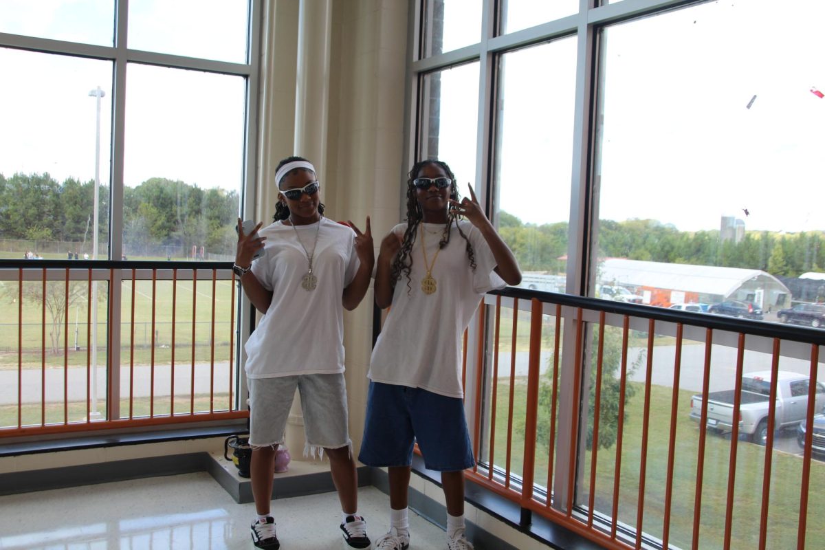 Two friends stand in the stairwell getting photos and videos of them dressed up to help remember the day. Dressed in white t-shirts, long jean shorts, sunglasses, gold chains, and bulky sneakers they are certainly the most fashionable of the day. 
