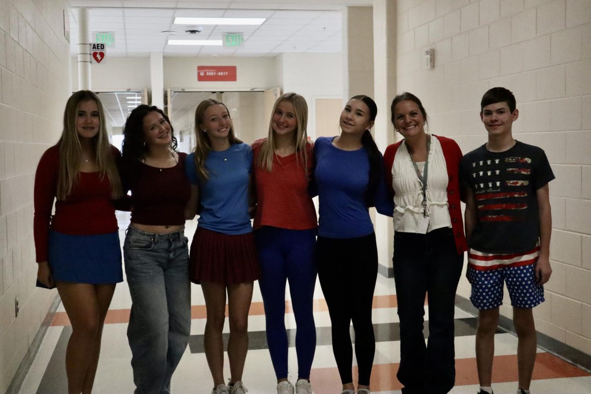 A group of students and their teacher stand in the hallway with their arms wrapped around each other.