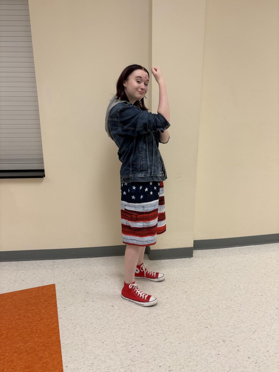 Junior Kate Price stands posing in a traditional rosie riveter pose in her jean jacket, red converse, and american flag shorts. 